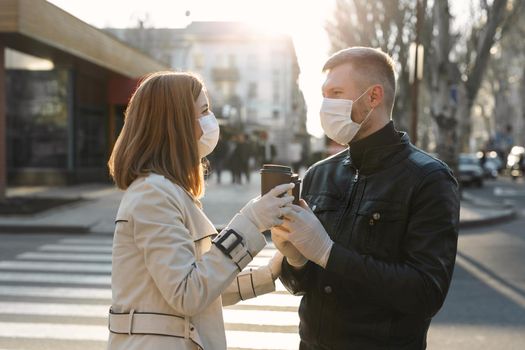 Young couple, a man and a woman in medical masks and gloves, drink coffee from disposable cups on the street and look at each other against. Coronavirus. Covid 19