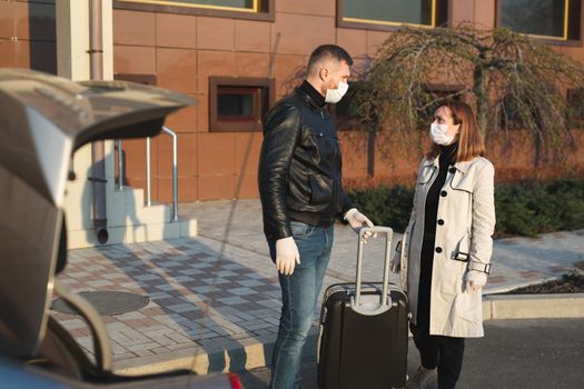 Man and woman in protective medical masks and gloves with a suitcase leave the house by car during the quarantine and self-isolation. The coronavirus. Covid 19.