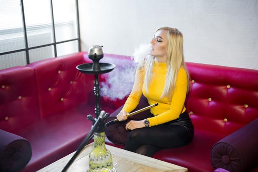 A beautiful young woman is sitting on a red leather sofa and Smoking a hookah.