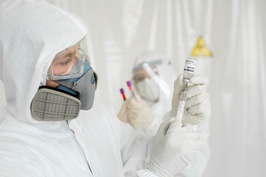 Doctor in a protective suit and mask holds an injection syringe and vaccine COVID-19. Laboratory Scientists in a Coverall Conducting a Research Vaccine.