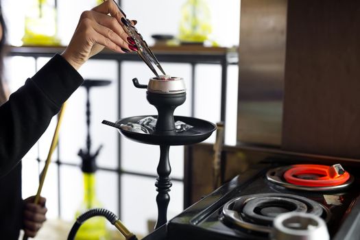 Women's hands hold hookah tongs and adjust the hot coals in a metal bowl. Black hookah stands in a restaurant or bar.