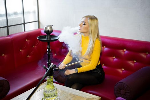 A beautiful young woman is sitting on a red leather sofa and Smoking a hookah.