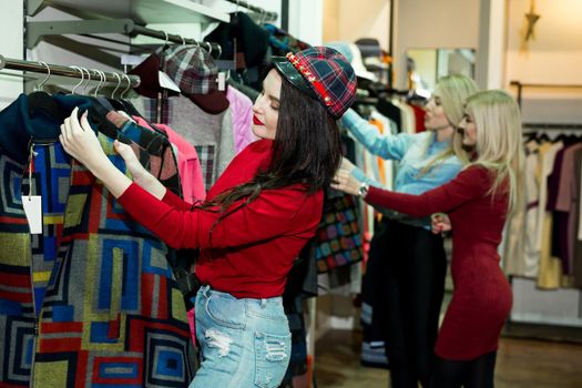 Three joyful young women shopping warm jackets at the apparel store. Shopping, fashion, clothes, style and people concept - happy woman trying coat on in mall or clothing store