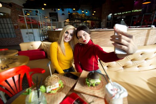 Two joyful cheerful girls taking a selfie while sitting together at cafe