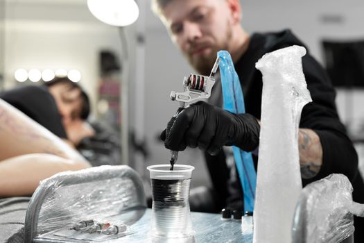 Tattoo master dips a tattoo machine with needle in water. Tattoo artist prepares tools and ink before work in tattoo parlor.