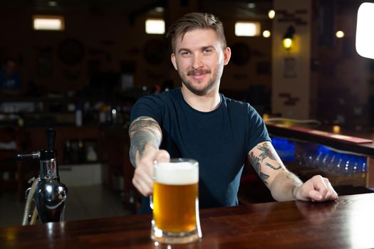 Handsome young male bartender in stretching out glass with beer and smiling while standing at the bar counter