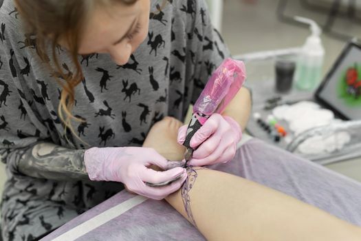 Master doing tattooing in tattoo studio. Professional tattooist works in studio. A woman in pink gloves makes a tattoo on the leg of a young girl