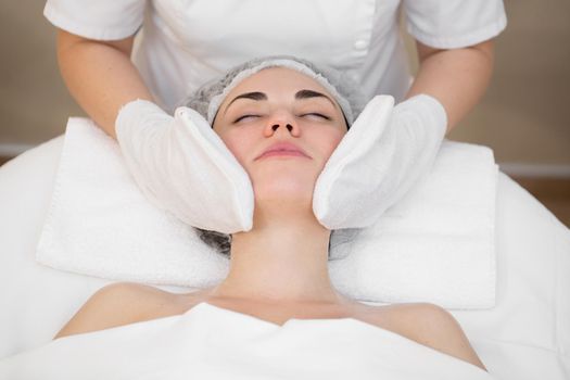 Hands of the beautician wipe the girl's face in the cosmetology center with white gloves. Portrait of a beautiful girl on the procedure for rejuvenating the face in a beauty salon