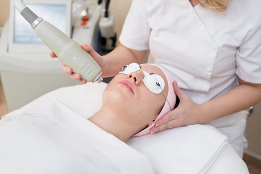 Young woman in safety glasses lying on the couch in a beauty salon, a beautician makes her facial rejuvenation procedure. Close-up of a procedure to rejuvenate the face for a young girl