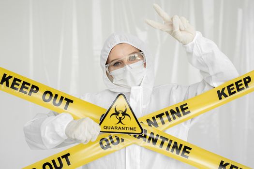 Epidemiologist woman in protective clothing is in a restricted area with a danger sign. Yellow line Keep Out Quarantine. Quarantine alert sign. Entrance is forbidden in quarantine zone.