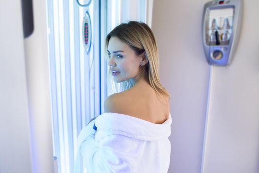 Sexy girl in a white robe is preparing for the tanning procedure in the Solarium. Vertical Solarium. Portrait of a slender girl in a modern Solarium