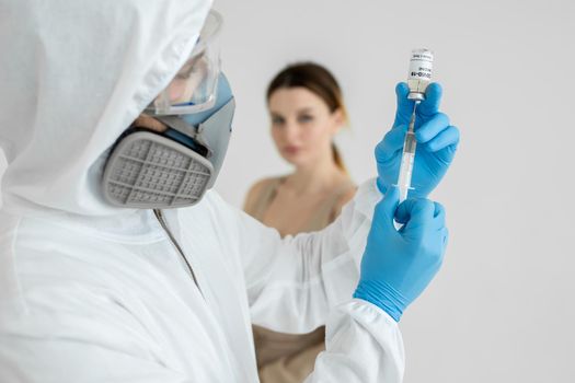 Doctor in a protective suit and mask holds an injection syringe and vaccine COVID-19. Man in a protective suit gives an injection to a woman from the coronavirus