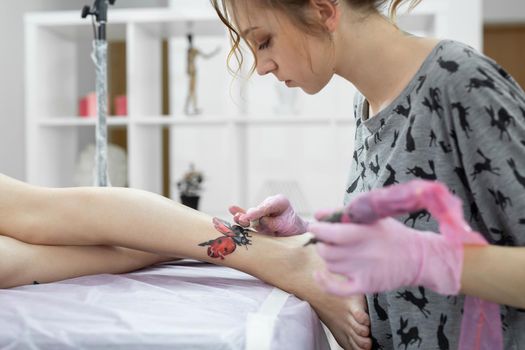 Master doing tattooing in tattoo studio. Professional tattooist works in studio. A woman in pink gloves makes a tattoo on the leg of a young girl