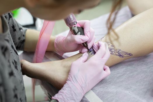 Close-up of a tattoo artist doing a tattoo on the leg of a young girl in the salon. Professional tattooist doing tattooing in studio