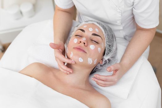 Hands of beautician apply cream to face of woman. Concept of beauty and youth. Close-up beautician causes moisturizer on the girl's face