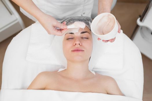 Face peeling mask, spa beauty treatment, skincare. Woman getting facial care by beautician at spa salon. Woman makes an alginate mask. Girl with a mask of beige clay