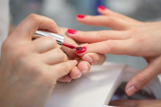 Woman nail master doing nails to a girl client at a beauty salon. A beautician applies red nail polish to a young womans nails