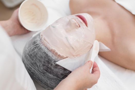 Face peeling mask, spa beauty treatment, skincare. Woman getting facial care by beautician at spa salon. Woman makes an alginate mask. Girl with a mask of beige clay
