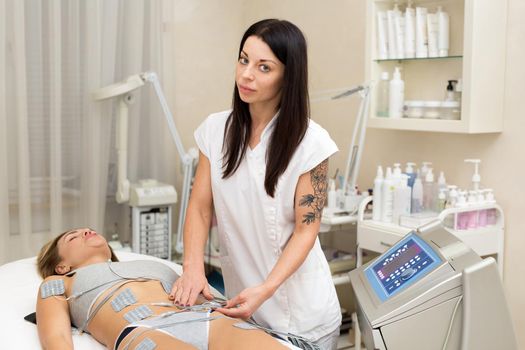 Professional beautician makes anti-cellulite massage to a young woman with the help of a biostimulating apparatus. Woman in beauty medical spa center anti-cellulite electrostimulation therapy