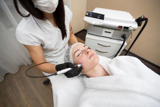 Therapist beautician makes a laser treatment to young woman's face at beauty SPA clinic. Close-up process of laser removal of blood vessels from the skin