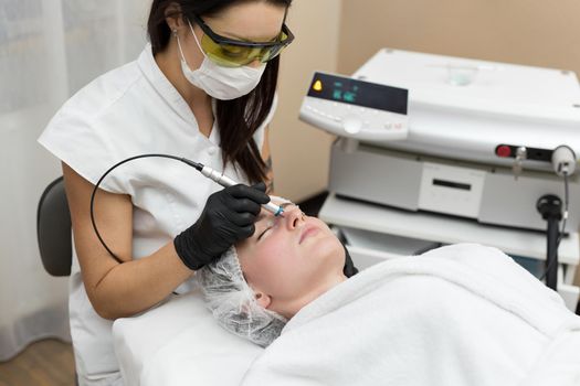 Cosmetologist in protective glasses conducts laser treatment of the skin for a young patient in a cosmetology clinic. Removal of vessels using a diode laser