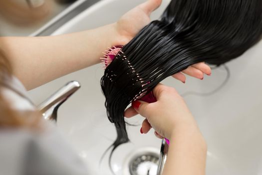 Young girl enjoys hair care in a beauty salon. The hairdresser washes the clients girls hair, applies a moisturizing oil mask and combs the hair with a comb. The process of hair care at hairdresser
