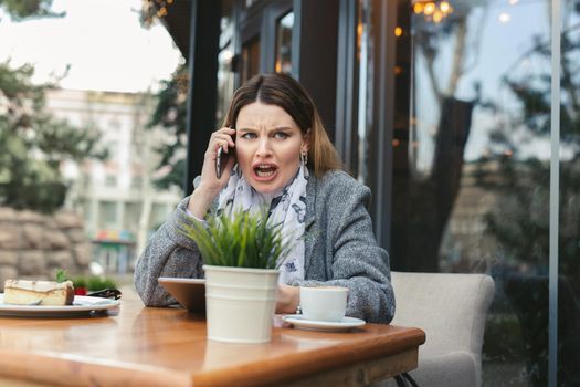 Portrait of angry young woman solving business problems shouting during phone conversation sitting in cafe terrace.Disappointed female upset about getting bad news from employee talking on cellular.