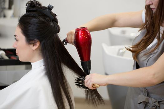 Hair stylist work on woman hairstyle in salon. Drying long brown hair with hair dryer and round brush