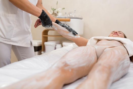 Beautiful sexy young woman receiving cosmetic clay, marine algae body mask lying on massage table at beauty salon. Close-up cosmetologist applies a moisturizing mask on the hand of a young woman.