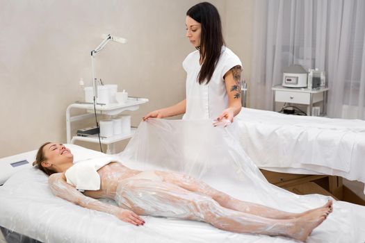 Cosmetic procedure wraps. The beautician covers the young girl with cellophane for anti-cellulite hydro-massage in a modern cosmetology room. Moisturizing body mask and body wrap.