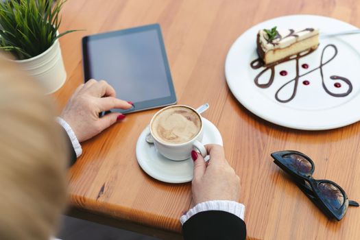 Woman drinks coffee in a cafe on the street. Close-up of a tablet, glasses, coffee and dessert on a wooden table.
