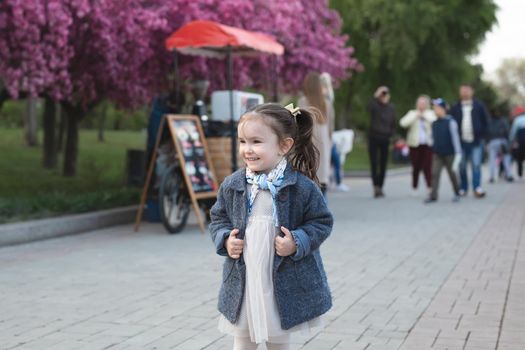 Little cute girl in a coat walks in the park against the background of a flowering tree in spring.