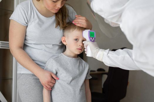 Pediatrician or doctor checks elementary age boy's body temperature using infrared forehead thermometer gun for virus symptom - epidemic coronavirus outbreak concept.
