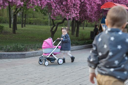 Little girl in a coat walks in the park and rolls a toy stroller with a doll.