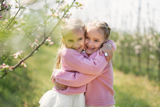 Happy two twin sisters are hugging against the background of a green blossoming apple orchard.