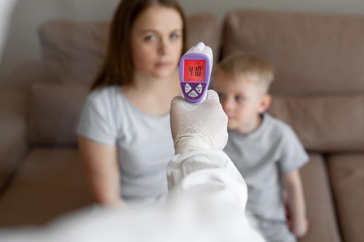 Doctor checks the body temperature of the girl and her son using an infrared thermometer gun on the forehead at home. Coronavirus, covid-19, high fever and cough.