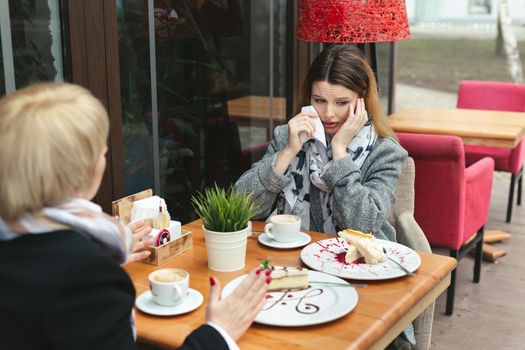 Sad and unhappy daughter sitting in cafe or restaurant with her mother, crying and wipes her tears with a paper napkin.