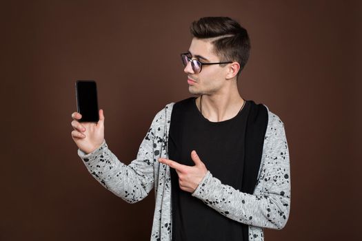 Close up portrait of young handsome male model showing smartphone to camera, isolated over brown background.