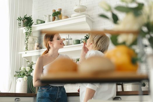 An adult daughter feeds her elderly mother fresh pastries in the kitchen.