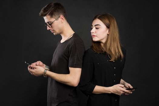 Profile portrait of young married couple, browsing information at their pdas, standing back to back, wearing casual outfits on black background. Another life in social nets