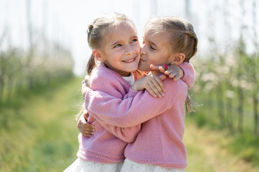 Happy twin sisters hug against the background of a green blooming Apple orchard. One sister kisses the other on the cheek.