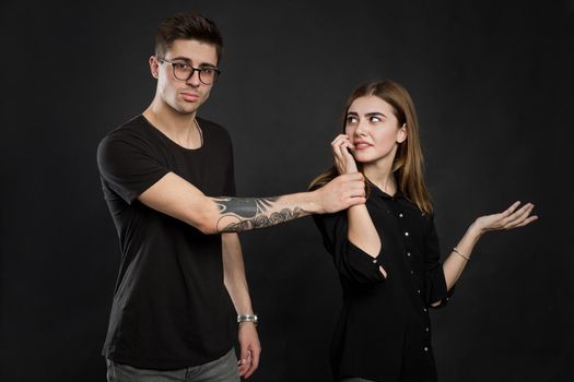 Portrait of a young couple standing with mobile phone, girl holding mobile phone while frustrated man standing near isolated over black background