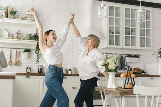 Mother and her adult daughter are having fun and dancing in the kitchen.