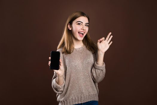 Smiling woman is pointing on smartphone standing on brown background