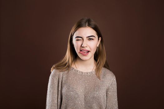 Cheerful young beautiful girl smiling showing tongue looking at camera over brown background