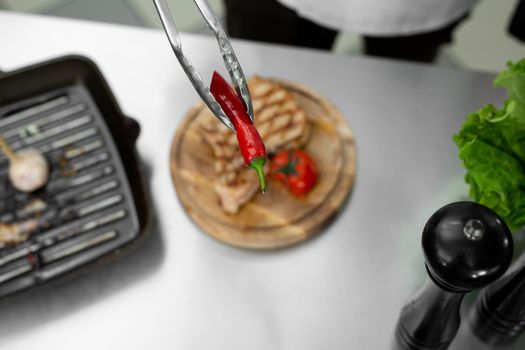 Close-up of the chef's hand as he puts chili peppers from the grill on a plate with a pair of tongs.