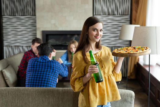 Young beautiful girl poses with beer and pizza in a cafe against the background of her friends.