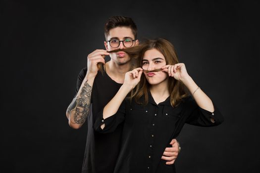 Beautiful young loving couple making fake moustache from hair while standing against black background. Funny moustache.