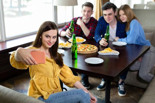 Group or company of young people - friends drink beer, eat pizza, talk and laugh and shoot selfie on the smartphone's camera on the background of the bar.