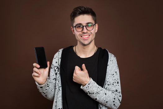 Close up portrait of young handsome male model showing smartphone to camera, isolated over brown background.